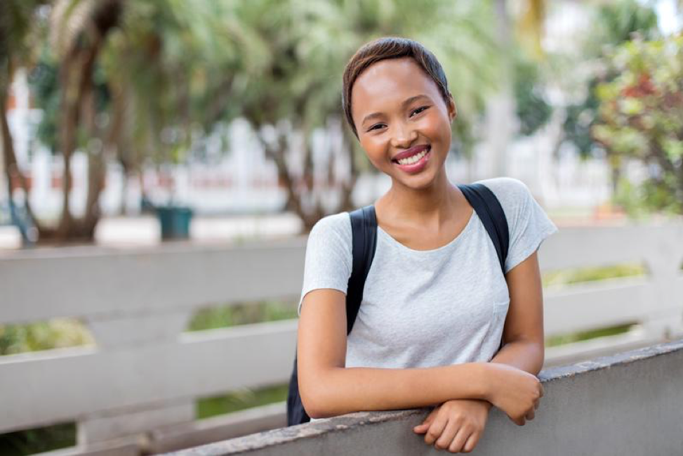 A young woman with a backpack is leaning on a fence and smiling.